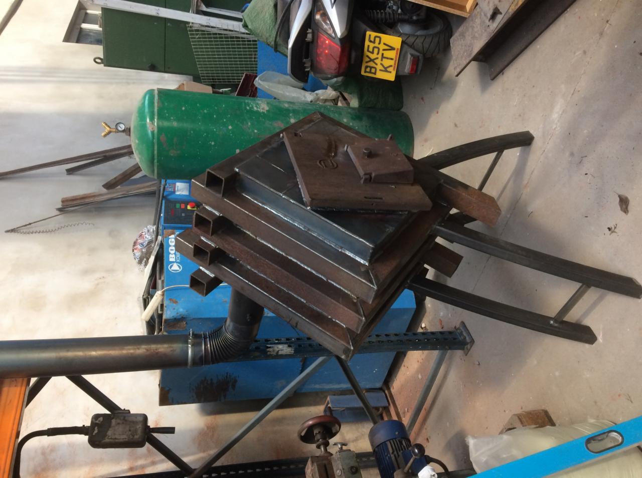 Stos kwadratowych, metalowych elementów, prawdopodobnie pokryw lub płyt, ułożonych na metalowej konstrukcji w warsztacie, obok spawarki i butli z gazem, z motocyklem w tle.