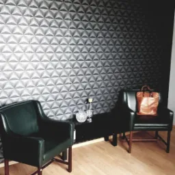 Dwa zielone fotele klubowe z brązowymi drewnianymi nogami na jasnej podłodze, czarny stolik z karafką i szklankami, torebka na fotelu, ściana z trójwymiarową tapetą.