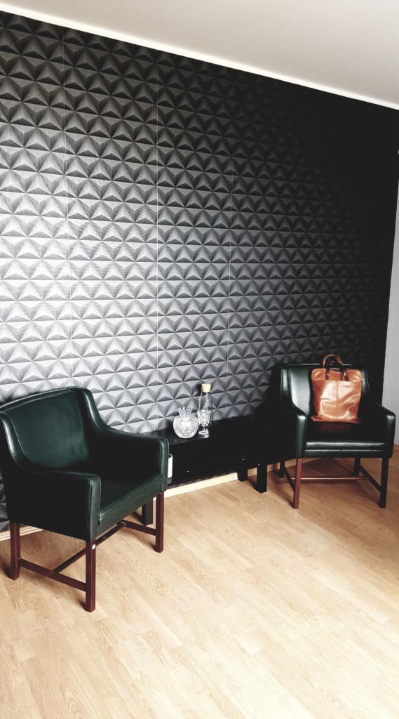 Dwa zielone fotele klubowe z brązowymi drewnianymi nogami na jasnej podłodze, czarny stolik z karafką i szklankami, torebka na fotelu, ściana z trójwymiarową tapetą.