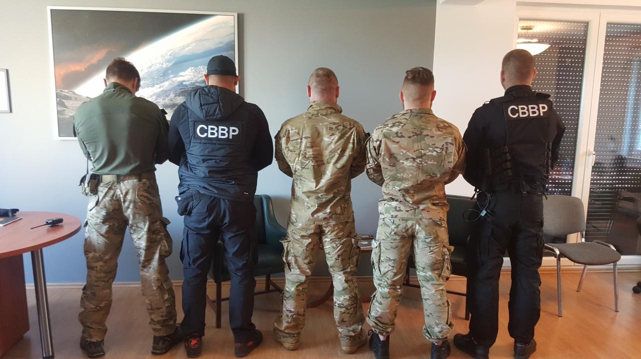 Pięciu mężczyzn w mundurach stojących tyłem, z napisem 'CBBP' na niektórych ubraniach, w pomieszczeniu z biurkiem i obrazem kosmosu na ścianie.