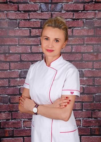 Portret kobiety w białym uniformie z różowymi akcentami, skrzyżowane ręce, zegarek na nadgarstku, na tle ściany z cegły.