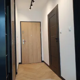 Korytarz z ułożoną parkietową podłogą w jodełkę, widoczne dwie drewniane drzwi z czarnymi ościeżnicami i listwami przypodłogowymi, na suficie czarna listwa z reflektorami, ściany pomalowane...