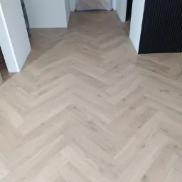 Ułożone panele podłogowe w jodełkę francuską w jasnym odcieniu drewna, widoczne białe futryny drzwi i czarna ściana z pionowymi panelami dekoracyjnymi.