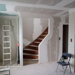 Wnętrze w trakcie prac wykończeniowych: ściany z płyt gipsowo-kartonowych, widoczne schody drewniane, drabina oparta o ścianę, krzesło z butelką. Surowy stan, przygotowanie do malowania.