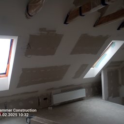 Poddasze w trakcie prac wykończeniowych: okna dachowe, ściany przygotowane pod gładzie, grzejnik. Widoczne zabezpieczenia belek stropowych folią.