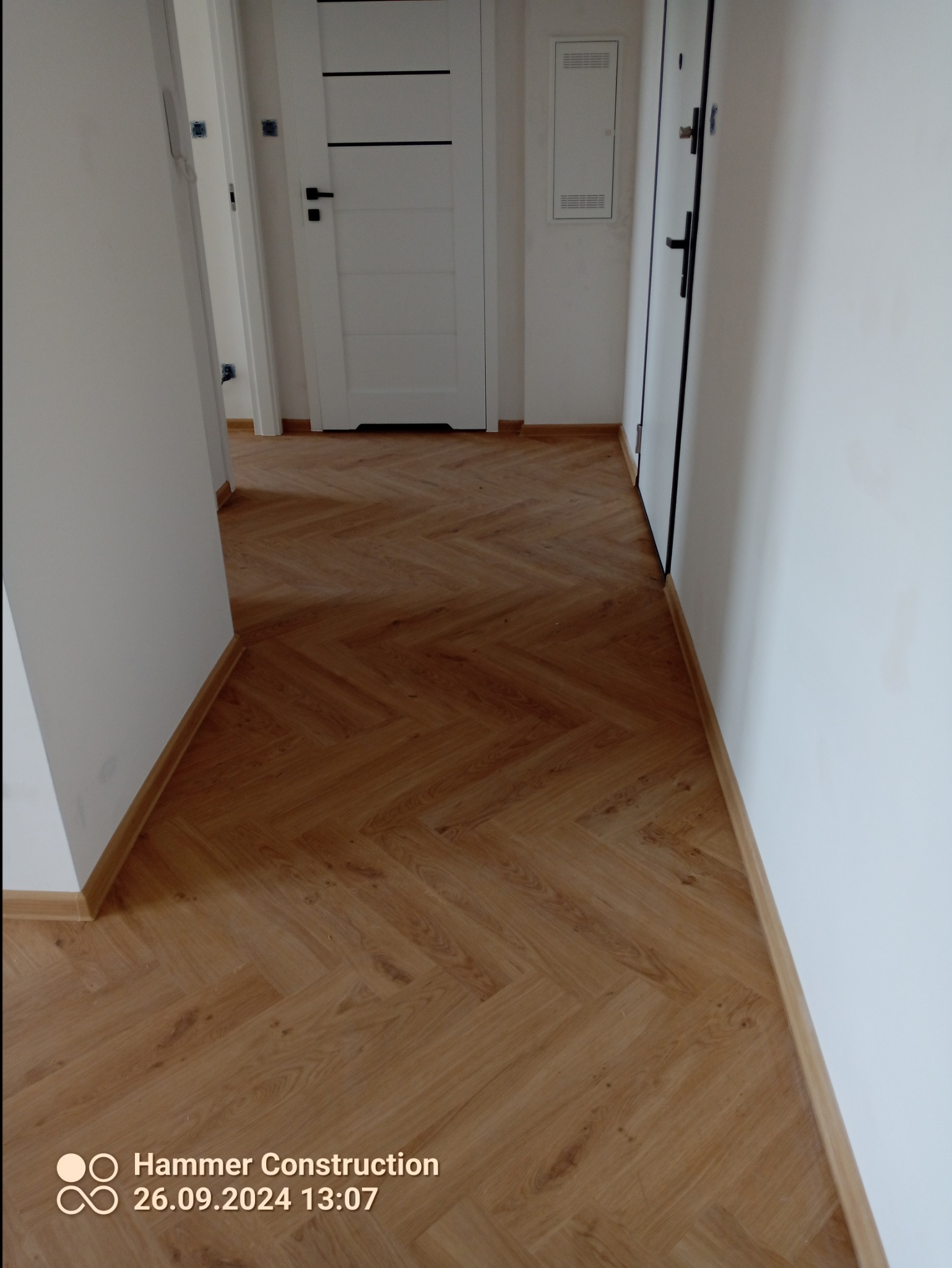 Ułożona podłoga z paneli w jodełkę w korytarzu z białymi ścianami i drzwiami.