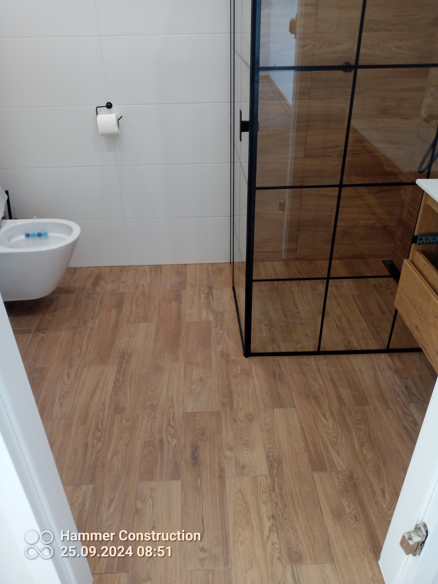 Nowoczesna łazienka z drewnopodobną podłogą, białą armaturą i kabiną prysznicową z czarnymi profilami, widok na bidet z niebieskimi kostkami odświeżającymi.