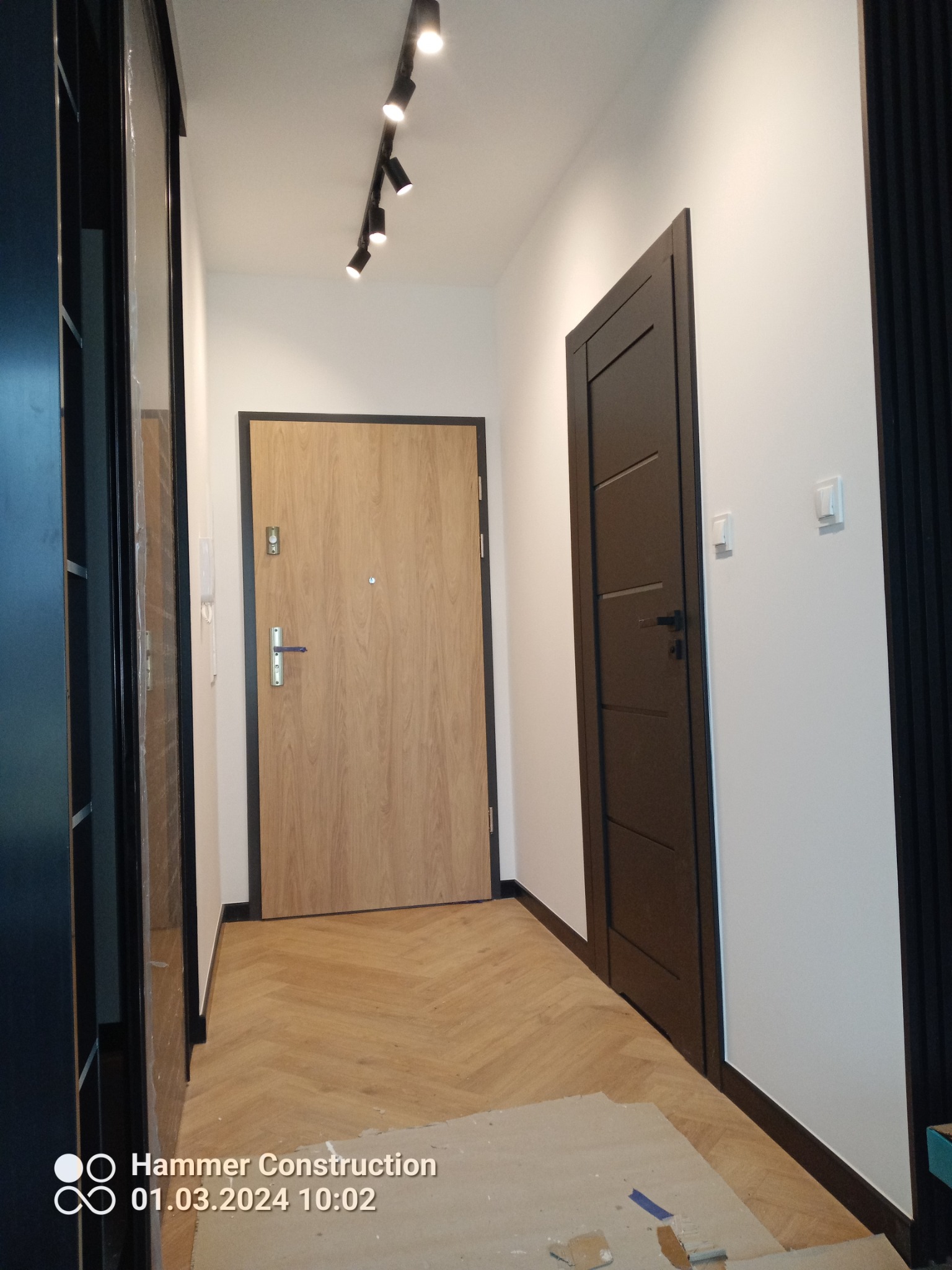 Korytarz z ułożoną parkietową podłogą w jodełkę, widoczne dwie drewniane drzwi z czarnymi ościeżnicami i listwami przypodłogowymi, na suficie czarna listwa z reflektorami, ściany pomalowane...