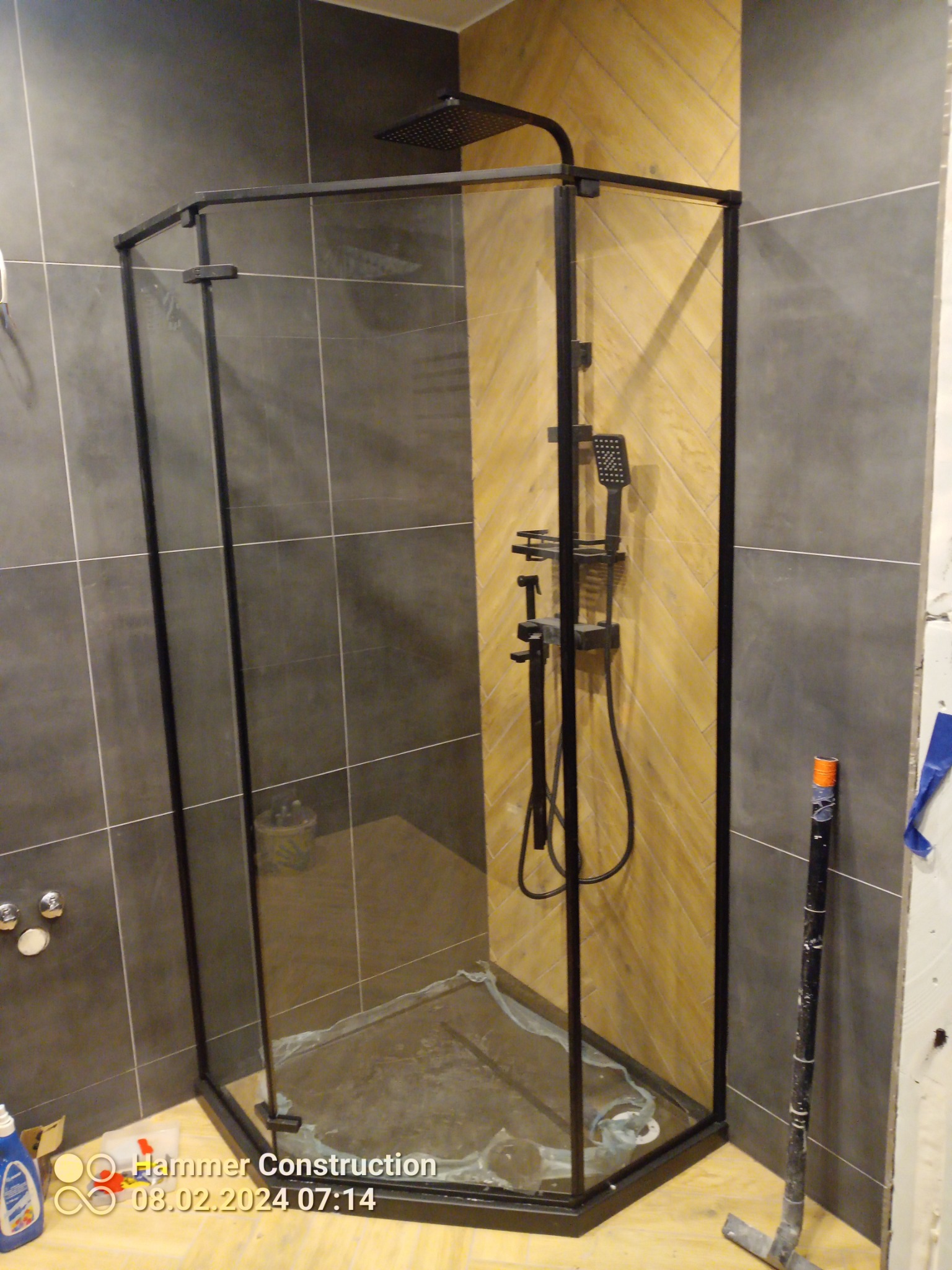 Nowoczesna kabina prysznicowa z czarnymi profilami, zamontowana w łazience z szarymi płytkami i ścianą wyłożoną drewnopodobnymi panelami w jodełkę. Wewnątrz kabiny widoczne pozostałości po pracach...