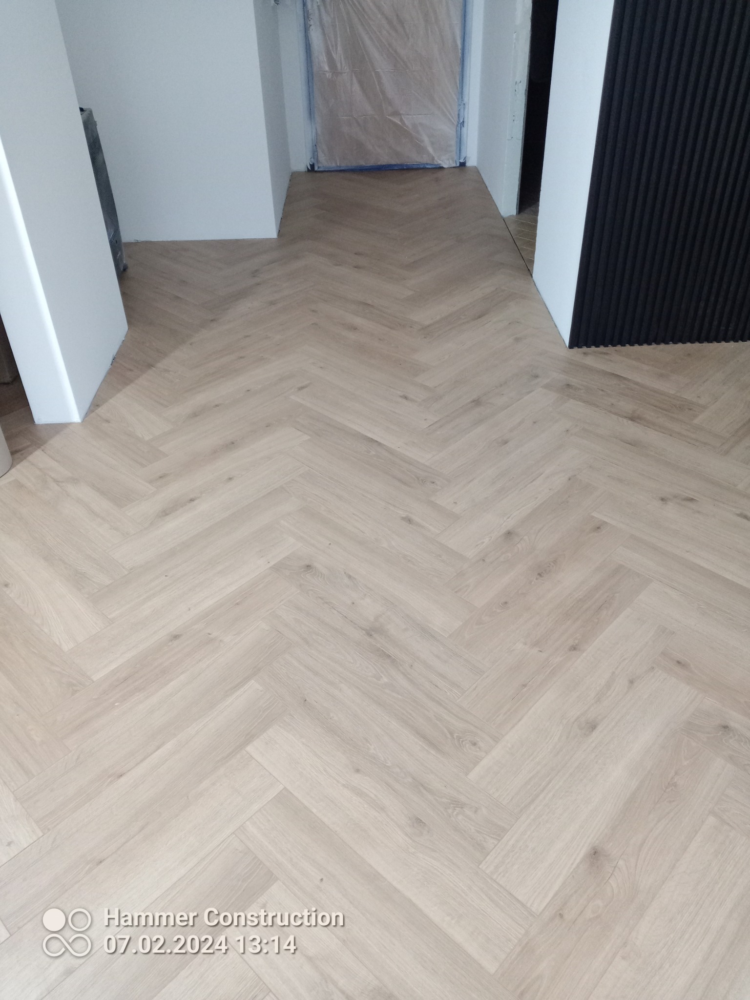 Ułożone panele podłogowe w jodełkę francuską w jasnym odcieniu drewna, widoczne białe futryny drzwi i czarna ściana z pionowymi panelami dekoracyjnymi.