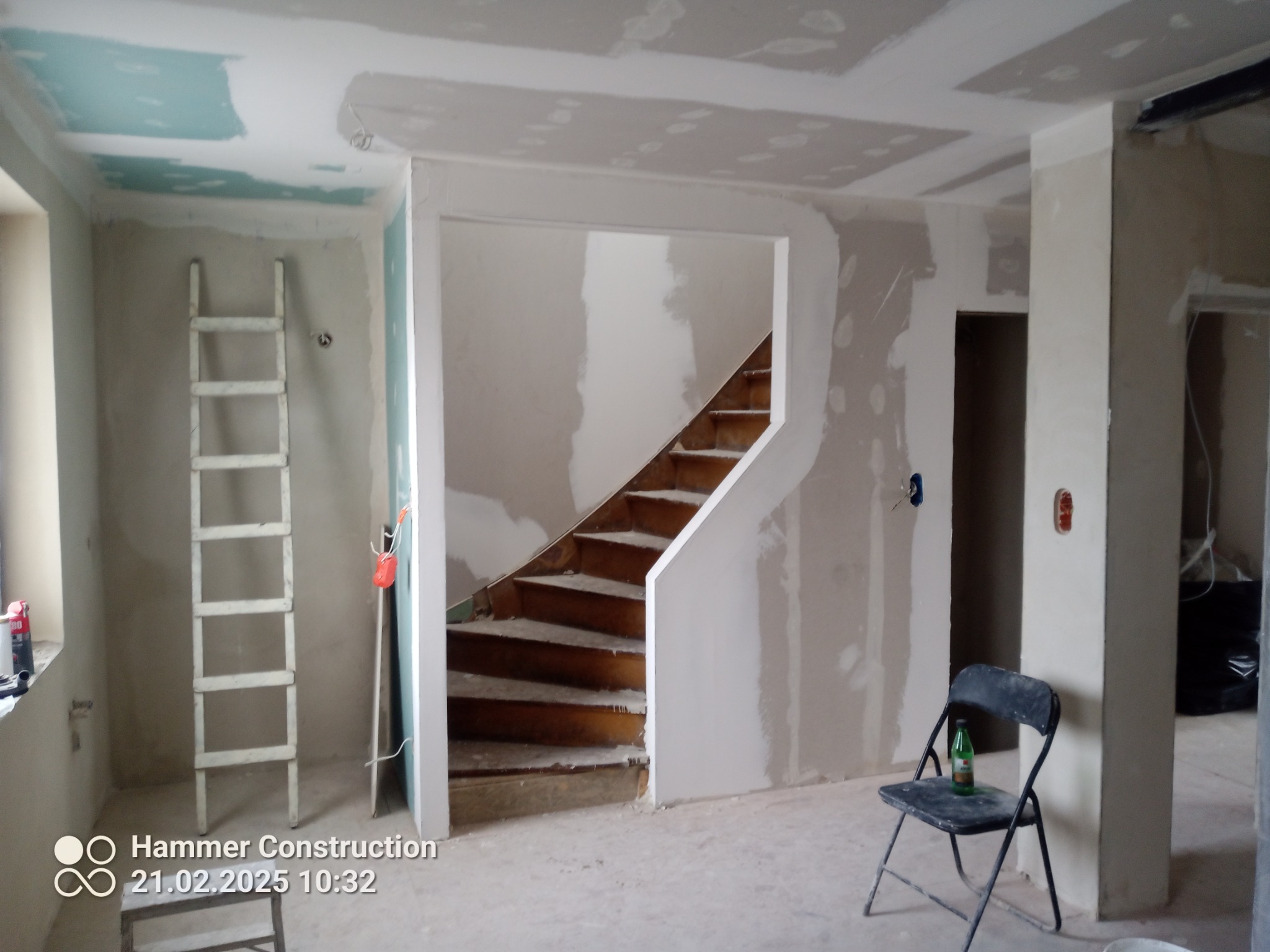 Wnętrze w trakcie prac wykończeniowych: ściany z płyt gipsowo-kartonowych, widoczne schody drewniane, drabina oparta o ścianę, krzesło z butelką. Surowy stan, przygotowanie do malowania.
