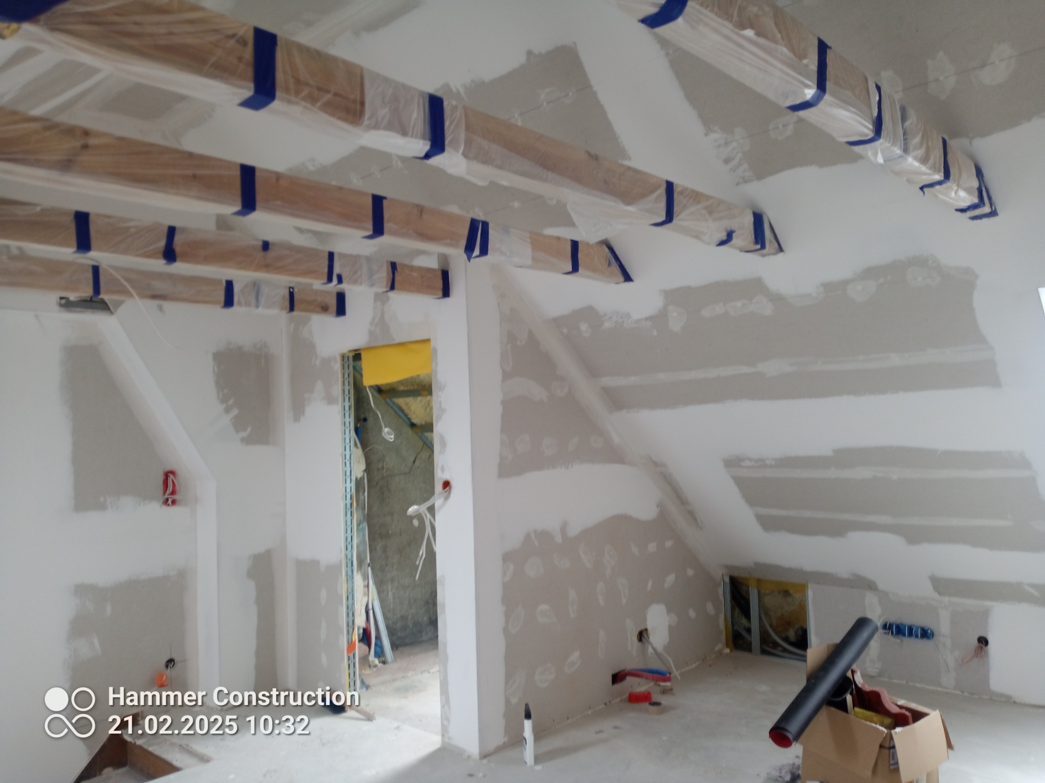 Poddasze w trakcie prac wykończeniowych: ściany z płyt gipsowo-kartonowych po szpachlowaniu, zabezpieczone folią drewniane belki stropowe, widoczne narzędzia i materiały budowlane.