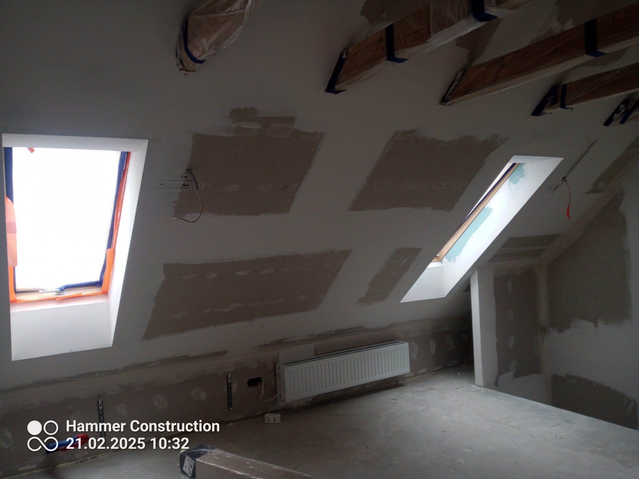 Poddasze w trakcie prac wykończeniowych: okna dachowe, ściany przygotowane pod gładzie, grzejnik. Widoczne zabezpieczenia belek stropowych folią.