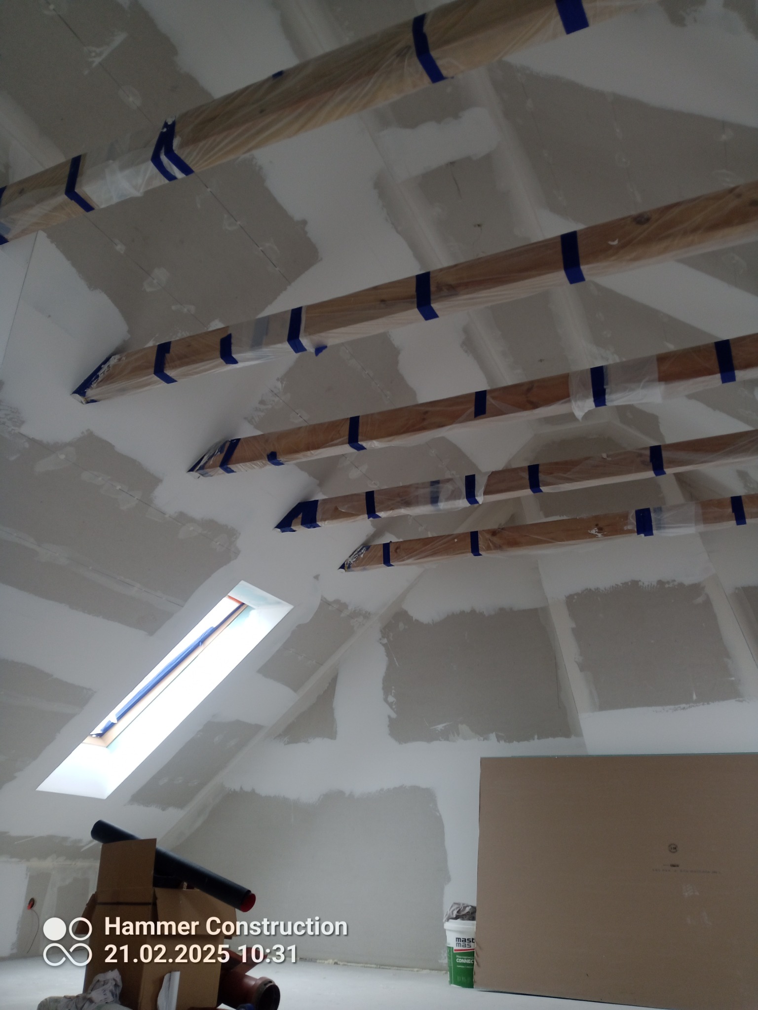 Poddasze z oknem dachowym w trakcie prac wykończeniowych. Widoczne drewniane belki zabezpieczone folią i taśmą, ściany pokryte gładzią szpachlową.