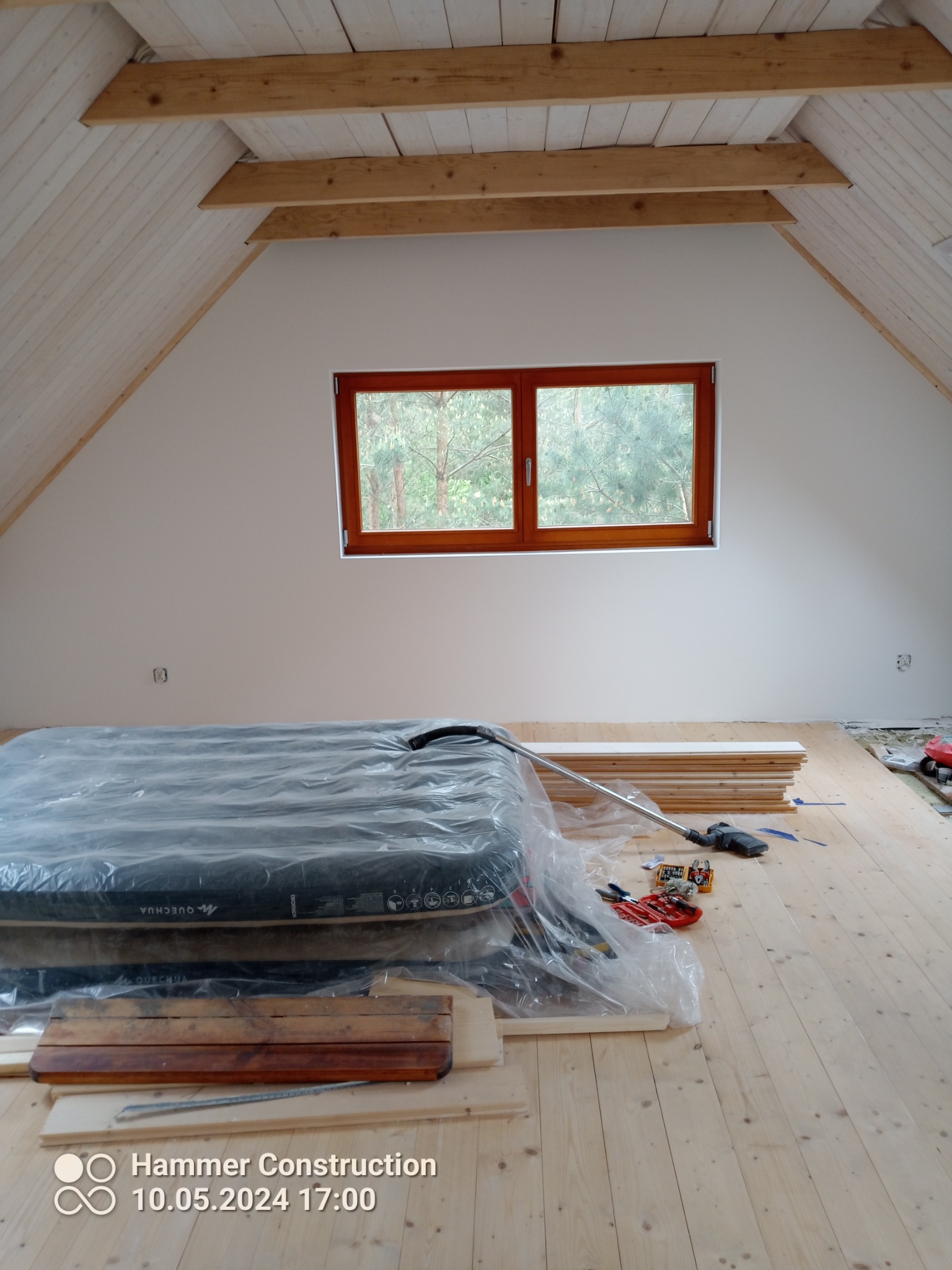 Poddasze z drewnianą podłogą w trakcie prac wykończeniowych. Widoczne deski, narzędzia i okno z widokiem na drzewa. Białe ściany i drewniane belki stropowe.