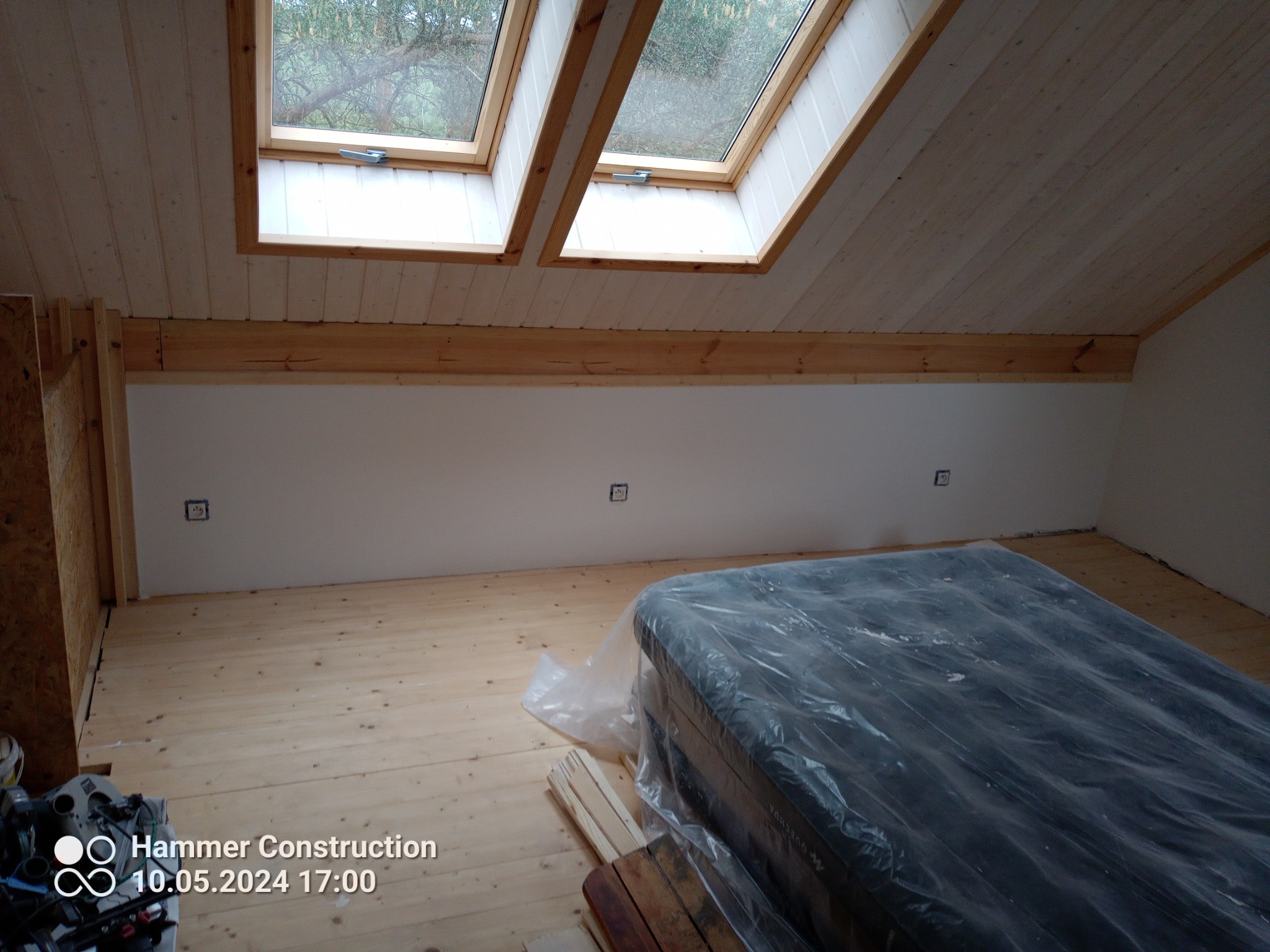 Poddasze z drewnianą boazerią, oknami dachowymi i jasną podłogą. Materac zabezpieczony folią na podłodze. Widoczne puszki elektryczne w ścianie. Etap prac wykończeniowych.
