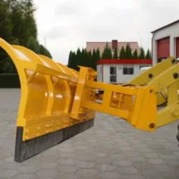 Żółty pług zamontowany na maszynie budowlanej, widoczne połączenia i elementy mocujące, w tle budynek z czerwonym dachem i zielone drzewa.