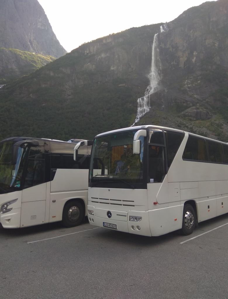 Dwa białe autokary Mercedes parkują na tle górskiego krajobrazu z wodospadem w Norwegii, widoczny numer rejestracyjny LU 476GY.