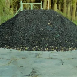 Kopiec ekogroszku na betonowym podłożu, widoczne pojedyncze, jasne bryłki, w tle drzewa i zielone krzewy.