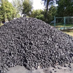 Góra czarnego węgla kamiennego na betonowym placu, w tle zielona siatka ogrodzeniowa i drzewa.