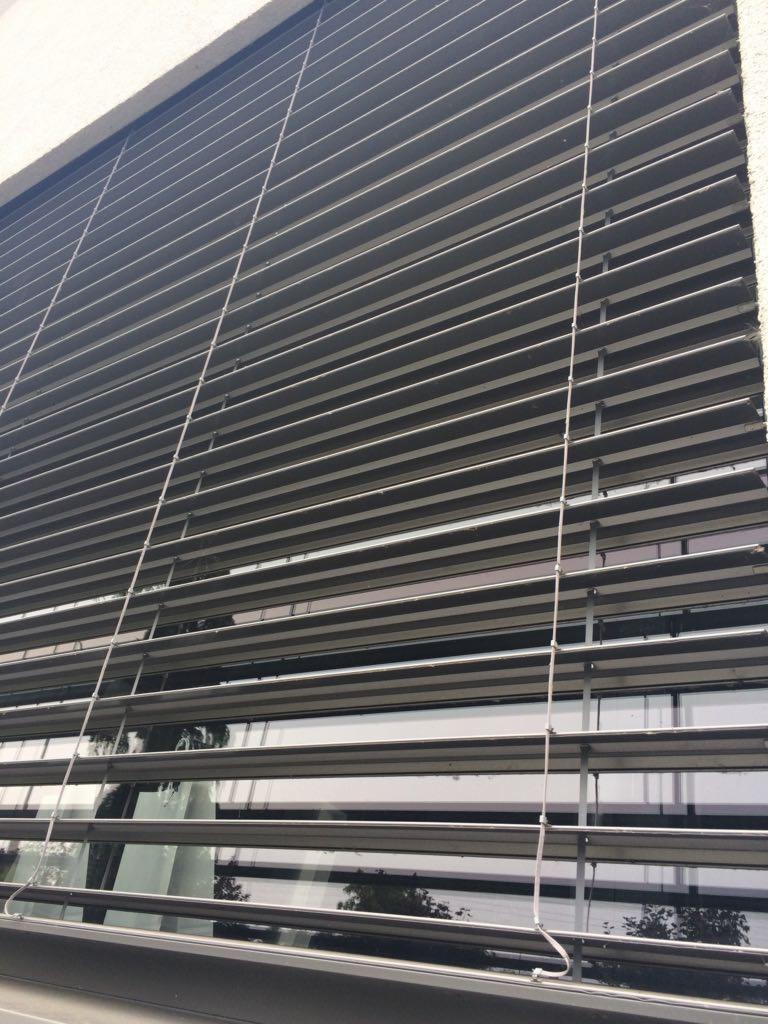 Zamknięte żaluzje aluminiowe w kolorze grafitowym, widok z dołu na okno, z widocznymi elementami konstrukcyjnymi i linkami sterującymi.