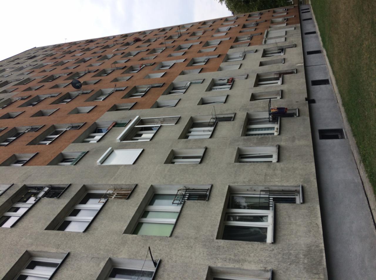 Wielopiętrowy budynek mieszkalny z widocznymi oknami, niektóre z balkonami, ujęcie z dołu w pochmurny dzień.