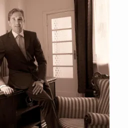 Portret mężczyzny w garniturze siedzącego na biurku w klasycznym gabinecie, z lampą na biurku i logo firmy widocznym w tle, utrzymany w sepii.