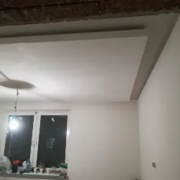 Nowo zamontowany biały sufit podwieszany z płyt kartonowo-gipsowych, widoczne niedoskonałości wykończenia przy ścianie, okno z widocznymi materiałami budowlanymi na parapecie.