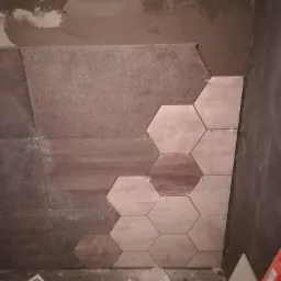 Montaż heksagonalnych płytek ściennych w odcieniach beżu i brązu, z widocznymi krzyżykami dystansowymi, na nierównej ścianie pokrytej szarym tynkiem.