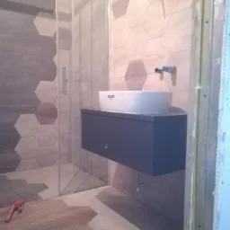 Nowoczesna łazienka w trakcie wykańczania: heksagonalne płytki na ścianach i podłodze, wisząca szafka z umywalką nablatową, widoczna kabina prysznicowa z przezroczystego szkła, czerwony klucz...