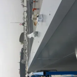 Szara, metalowa konstrukcja dachu hali w trakcie budowy, widoczne opakowania elementów montażowych i narzędzia na powierzchni dachu, w tle zabudowa miejska