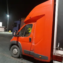 Pomarańczowy samochód dostawczy z plandeką na parkingu w nocy, widoczny fragment kabiny kierowcy.