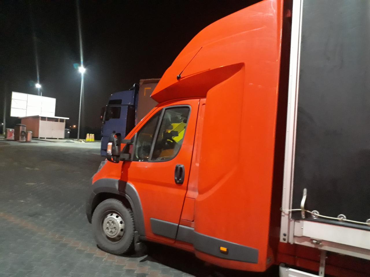 Pomarańczowy samochód dostawczy z plandeką na parkingu w nocy, widoczny fragment kabiny kierowcy.