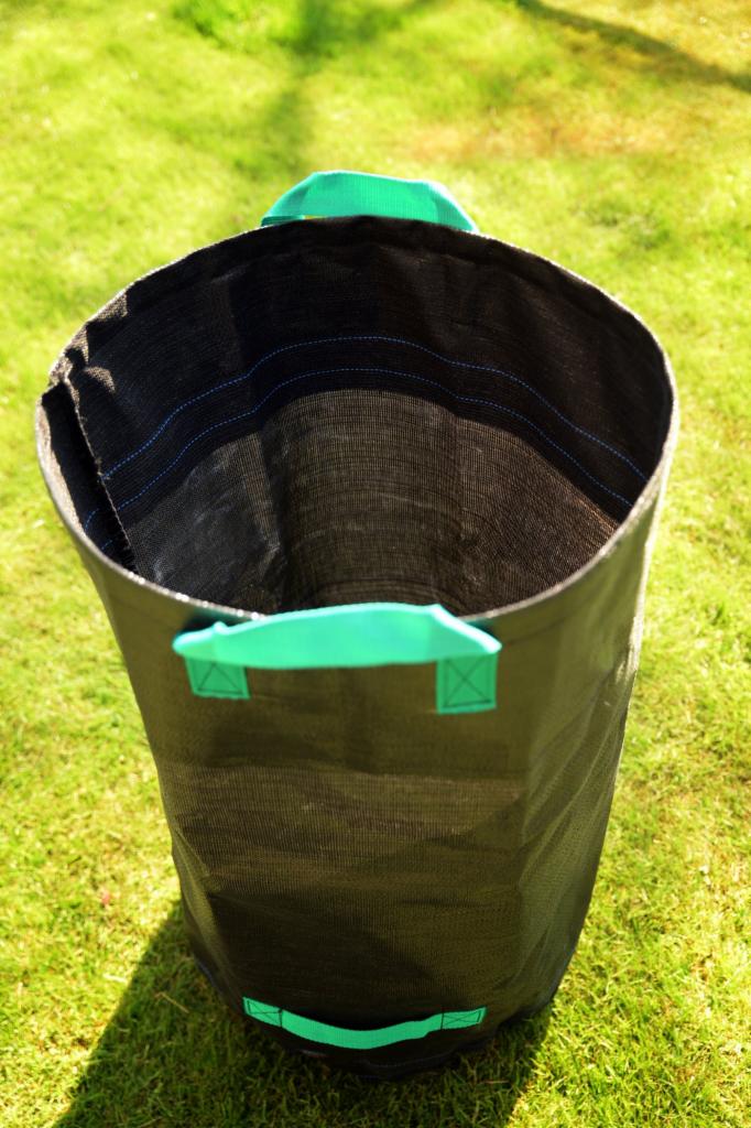 Czarny, tekstylny worek do uprawy roślin z zielonymi uchwytami, stojący na trawie w słoneczny dzień.