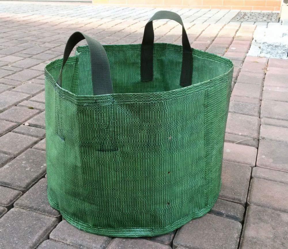 Zielony, tekstylny pojemnik na rośliny z uchwytami, stojący na kostce brukowej.