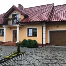 Żółta elewacja domu jednorodzinnego z czerwonym dachem, balkonem z kwiatami, kolumnami przy wejściu i ozdobnym podjazdem z kostki brukowej.