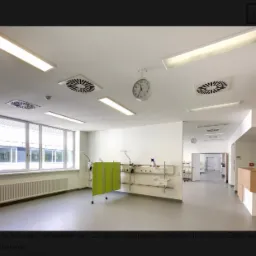 Jasne, przestronne wnętrze szpitala z białymi ścianami, szarym linoleum, dużymi oknami i zielonym parawanem medycznym. Na suficie widoczne lampy i kratki wentylacyjne, a na ścianie zegar.