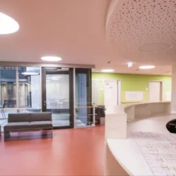 Jasne, nowoczesne wnętrze recepcji z elementami zieleni, szare sofy w strefie oczekiwania, widoczne plany architektoniczne na ladzie recepcyjnej.