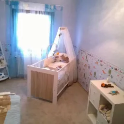 Pokój dziecięcy z łóżeczkiem, komodą i półką, ściany zdobione tapetą z motywami zwierzęcymi, niebieskie zasłony w oknie.