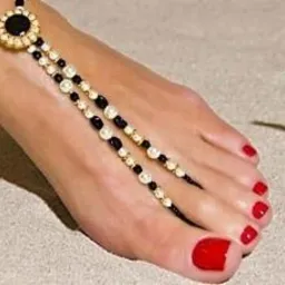 Pomalowane na czerwono paznokcie u stopy z ozdobną bransoletką na kostce, obok leży muszla na piasku.