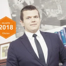 Portret mężczyzny w garniturze, z pomarańczową odznaką 'Najlepsi 2018 Oferteo', na tle regału z książkami i obrazu miasta.