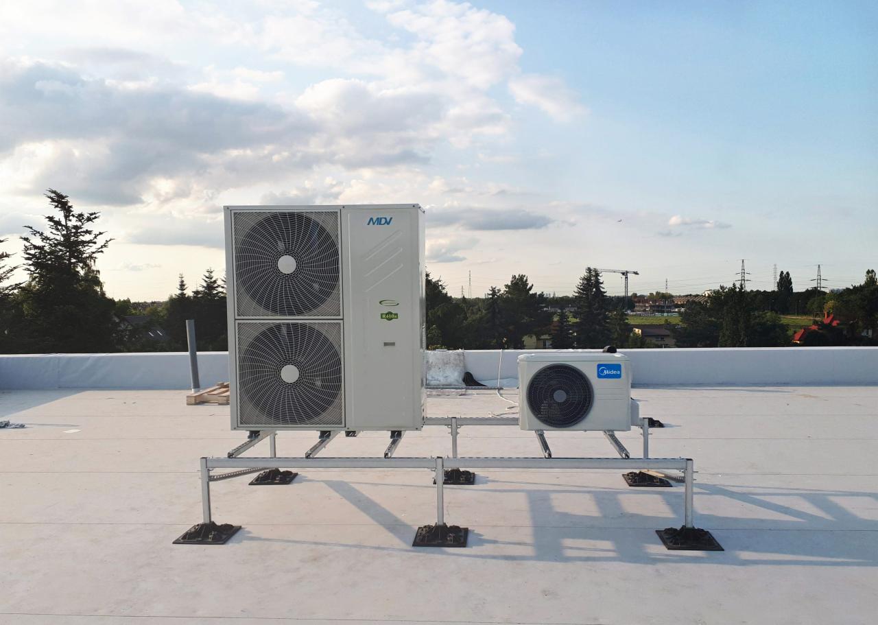 Dwa agregaty klimatyzacyjne MDV i Midea, zamontowane na metalowej konstrukcji na białym dachu budynku, widoczne drzewa i niebo w tle.