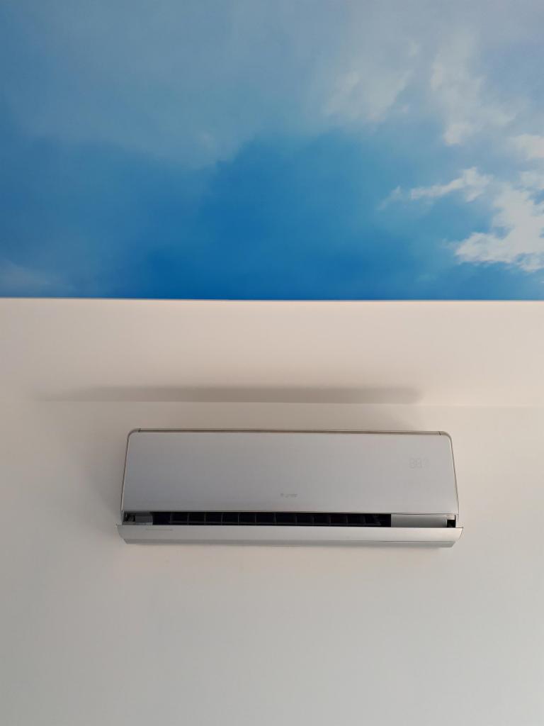 Nowoczesny, biały klimatyzator ścienny zamontowany na białej ścianie, z dekoracyjnym motywem nieba i chmur nad nim.