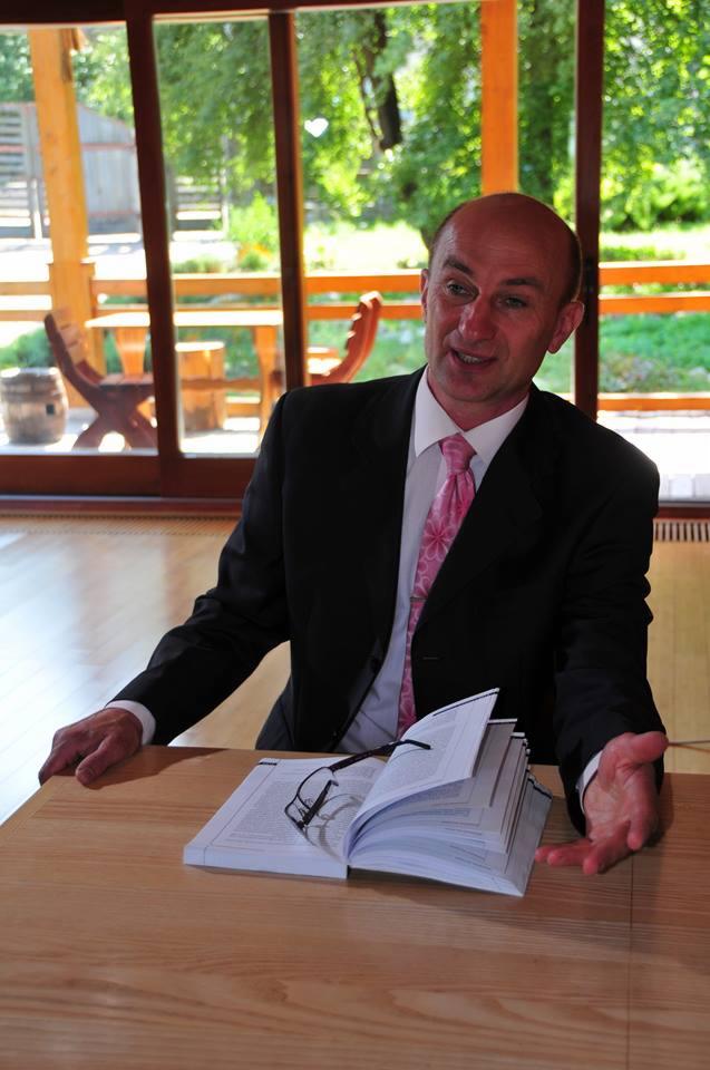 Mężczyzna w garniturze i różowym krawacie gestykuluje obok otwartej książki z okularami na drewnianym stole, w tle widoczne drewniane meble i zieleń.