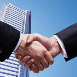 Uścisk dłoni dwóch osób w garniturach na tle wysokiego, nowoczesnego budynku biurowego i błękitnego nieba.