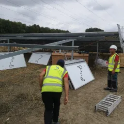 Montaż paneli słonecznych: dwóch pracowników w kamizelkach odblaskowych, panele leżące na ziemi, drabina, konstrukcja wsporcza w tle, pochmurne niebo.
