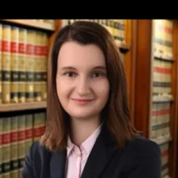 Portret uśmiechniętej kobiety w granatowej marynarce i różowej koszuli, na tle regału z książkami w kancelarii.