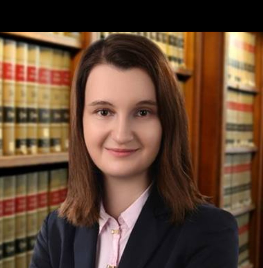 Portret uśmiechniętej kobiety w granatowej marynarce i różowej koszuli, na tle regału z książkami w kancelarii.