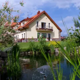 Jasny, dwupiętrowy budynek z czerwonym dachem i balkonem z kwiatami, otoczony zielenią i stawem z fontanną w słoneczny dzień.