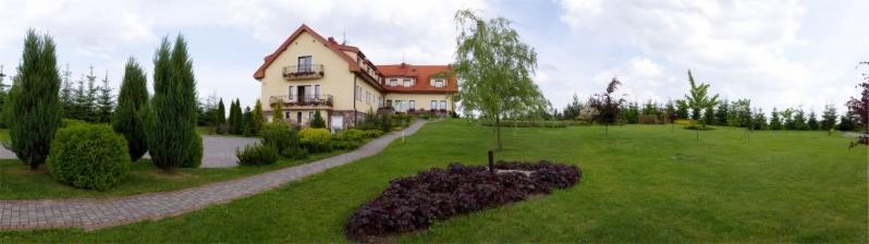 Panorama budynku kliniki z ogrodem, widoczne liczne drzewa iglaste, krzewy ozdobne i trawnik, ścieżka z kostki prowadząca do wejścia.